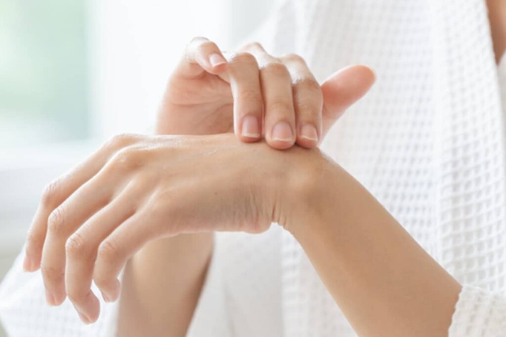 A woman in a white waffle-weave robe gently rubbing a topical cream into the back of her hand to moisturize and protect the skin.