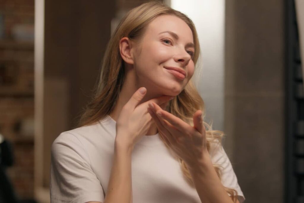 A woman with blonde hair smiling while gently touching her neck and jawline in a mirror to assess skin laxity and contour.