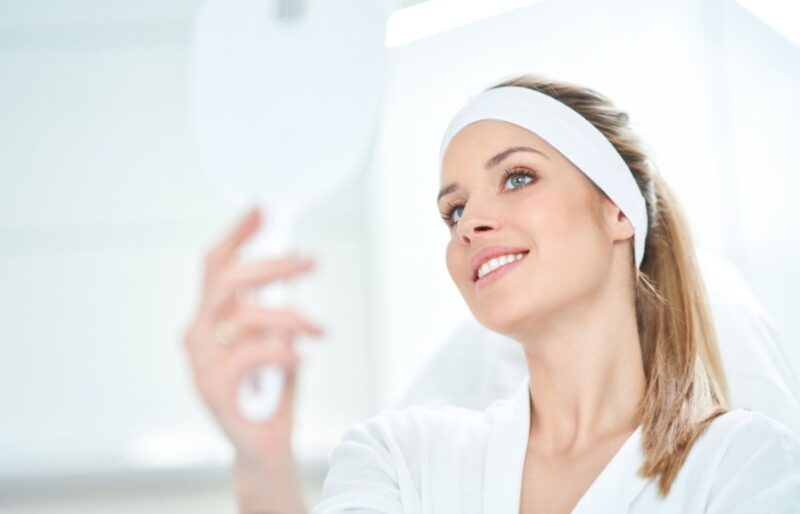 Woman examining smooth skin in mirror after Botox treatment, wearing headband and white robe in bright room.
