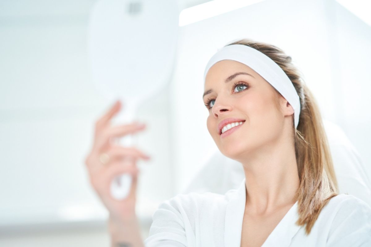 Woman examining smooth skin in mirror after Botox treatment, wearing headband and white robe in bright room.