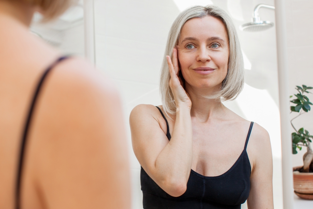Middle-aged woman examining her face in the mirror during facelift recovery week 3, showing defined facial contours.