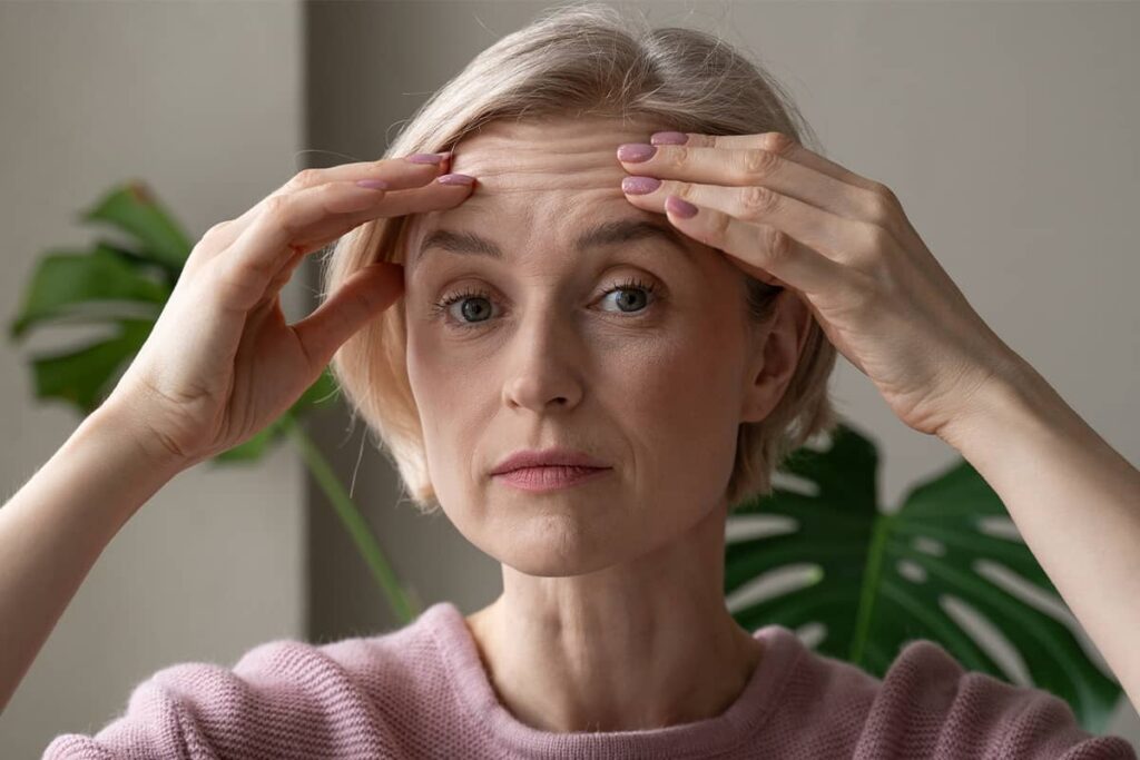 A woman with blonde hair looking into the camera while using her fingers to gently pull up and examine the horizontal frown lines on her forehead.
