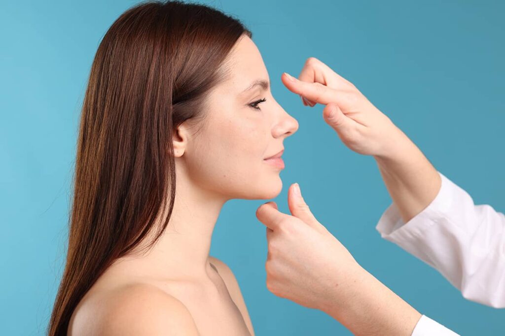 A profile view of a woman with long dark hair as a surgeon in a white coat assesses her nasal bridge and chin alignment against a blue background.